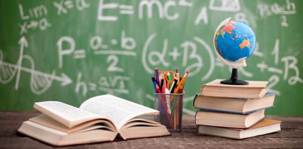 Materiais escolares sobre uma mesa, com livros abertos, globo terrestre e quadro negro ao fundo, representando a educação e o magistério.