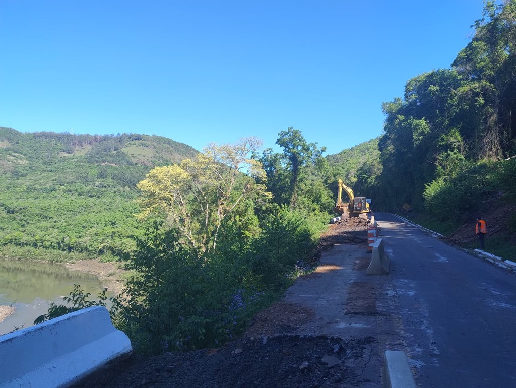 Escavadeira e operários atuam em obra de contenção na BR-116, em trecho de serra com vegetação densa e vista para o rio, entre São Marcos e Campestre da Serra.