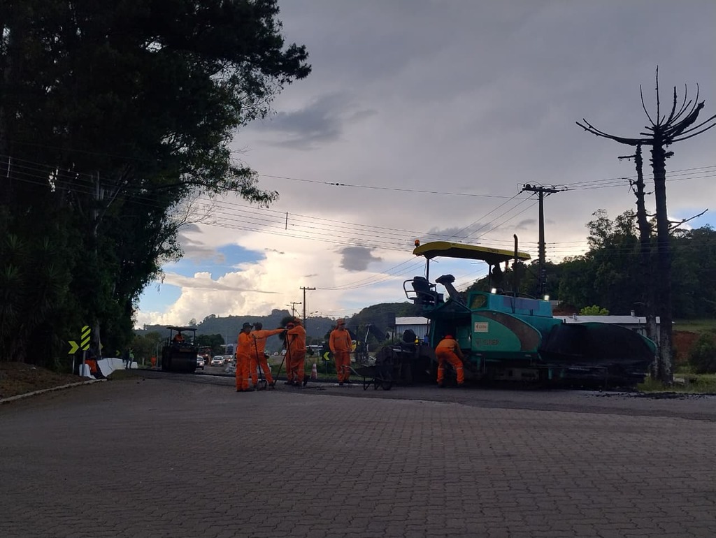 Equipe de operários trabalha em obra de pavimentação asfáltica na BR-116, utilizando máquina acabadora e rolo compactador, em trecho urbano de São Marcos.