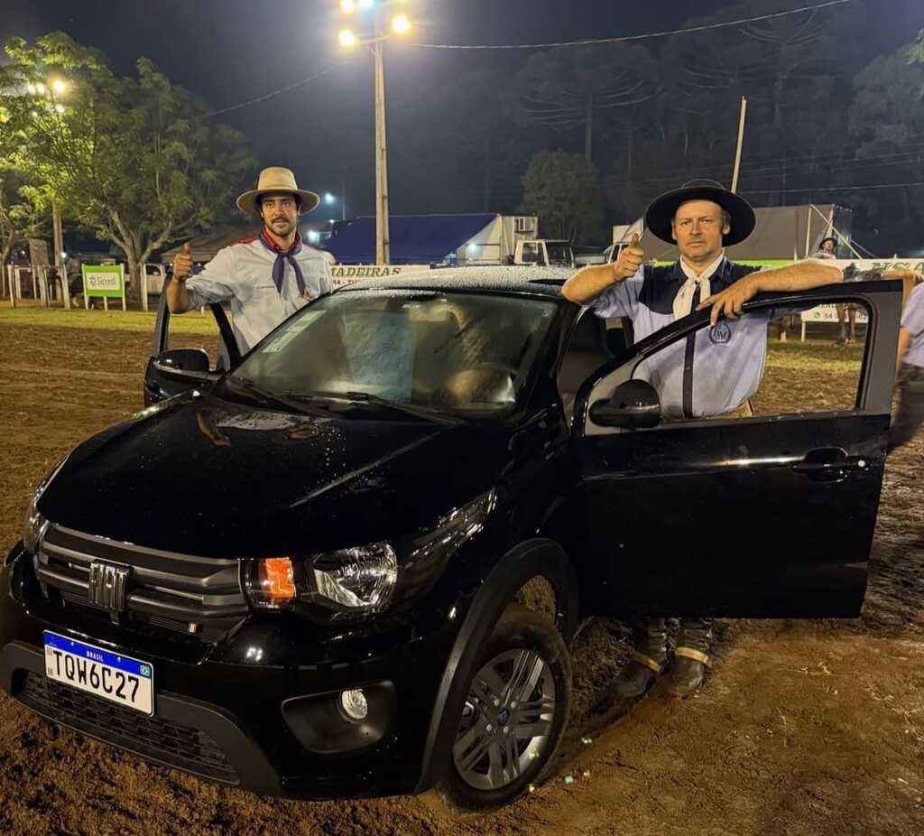 Dois homens pilchados, cada um ao lado de uma porta aberta de um Fiat Mobi preto, posam com expressão de comemoração após vencerem prova de laço. O carro está estacionado em uma cancha de rodeio à noite.