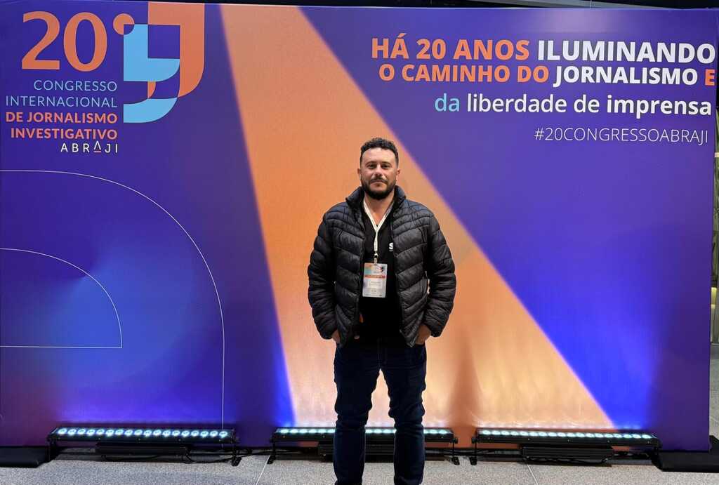 Angelo Batecini, fundador do São Marcos Online, em pé diante do painel oficial do 20º Congresso Internacional de Jornalismo Investigativo da Abraji, com credencial no peito e iluminação em tons de azul e laranja ao fundo.