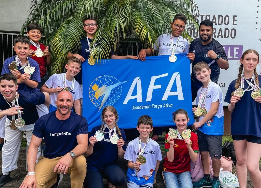 Grupo de jovens atletas da AFA Taekwondo posa sorridente ao ar livre, segurando diversas medalhas no peito. Eles exibem uma grande bandeira azul com o logotipo da AFA – Academia Força Ativa. Dois treinadores, um agachado na frente e outro em pé ao fundo, também posam junto ao grupo.