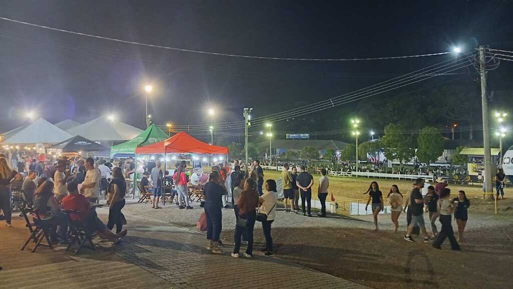 Vista noturna do Parque Municipal de Eventos durante o Rodeio de São Marcos, com grande circulação de público, tendas de alimentação iluminadas e a cancha de laço ao fundo, evidenciando a intensa movimentação do evento.